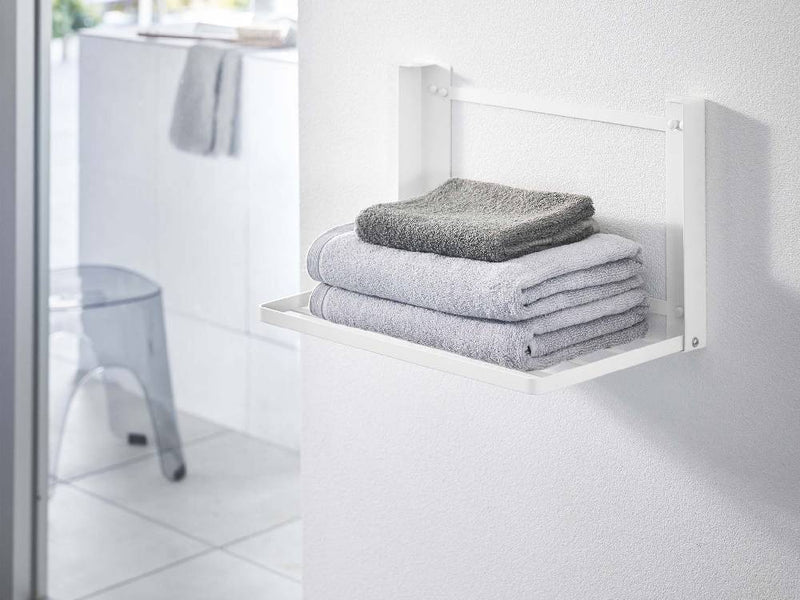 スチール製のタワー折り畳み棚が浴室の石こうボード壁に取り付けられ、タオルやバス用品が整然と収納されている様子