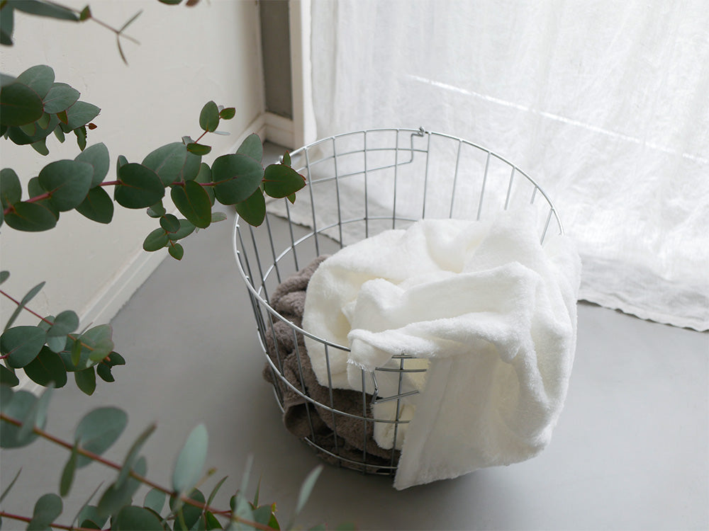 ナチュラル素材の柳編み収納バスケットにCUOLクオルのスキンケアタオルが美しく配置され、観葉植物やクッションと共に整えられた家庭用品のディスプレイ