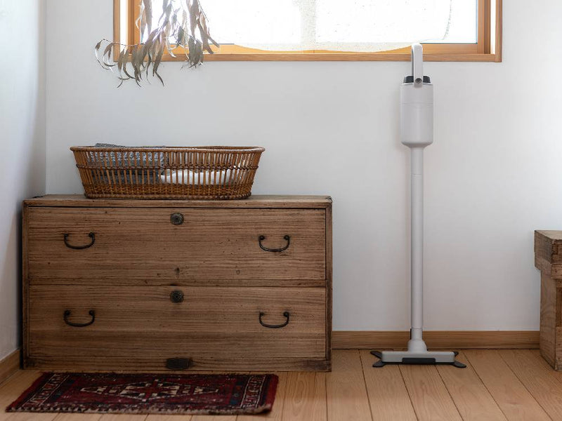 木製の床の上に置かれたプラスマイナスゼロ コードレスクリーナー G040。近くにランプと観葉植物が配置されています。