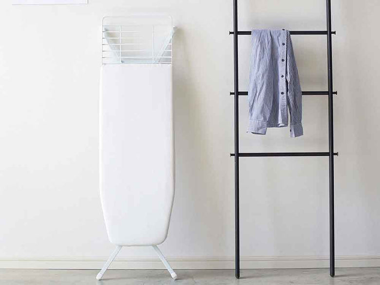 灰色のスタンド式アイロン台と衣類ハンガーが棚に配置された家庭用洗濯スペース