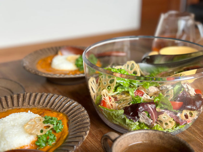 OXO/オクソーのクリアサラダスピナー小型モデルが透明なボウルとステンレス製フタで、野菜と一緒にテーブル上に配置されている様子