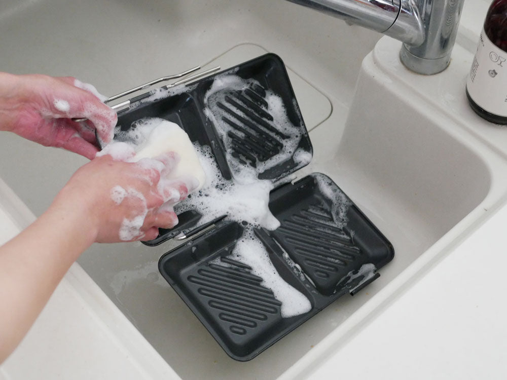 シンク横に配置された【下村企販】グリルdeクック ホットサンドパンとプラスチック製トレーに盛られた調理済み食品が写ったキッチン用品の画像