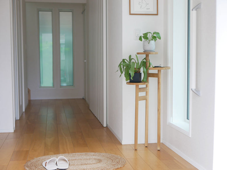 ナチュラルチーク無垢材で仕上げられた「サンフラワーラタン」ステップトレイテーブルが木製フローリングの室内に設置され、花鉢と共にインテリアとして飾られています