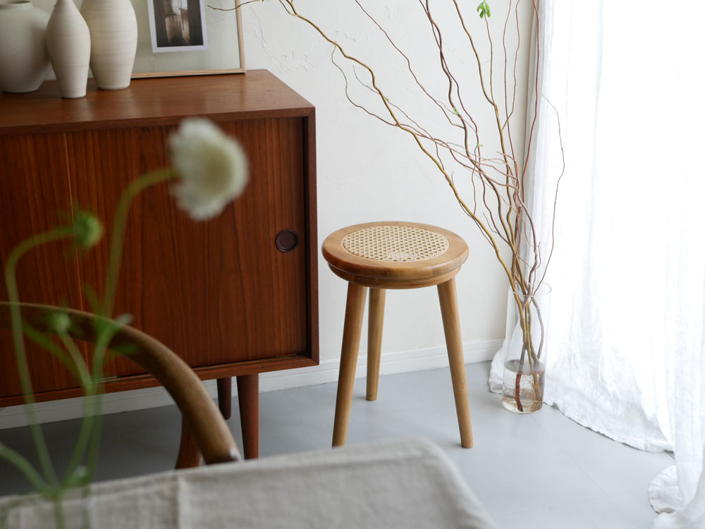 チーク材とラタンで作られたインドネシア産のサンフラワーラタンラタンスツールが木製の室内空間に置かれ、隣に花鉢が配置されている
