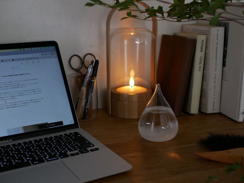 ガラス製のシリンダー型キャンドルランプが炎を灯し、木製ベースと観葉植物が配置されたオフィス用の静物写真