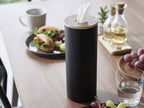 スチールと木材で作られたボトル型ティッシュケース Lが、カップやスプーン、ストローなどの食器と共にテーブルに配置されている
