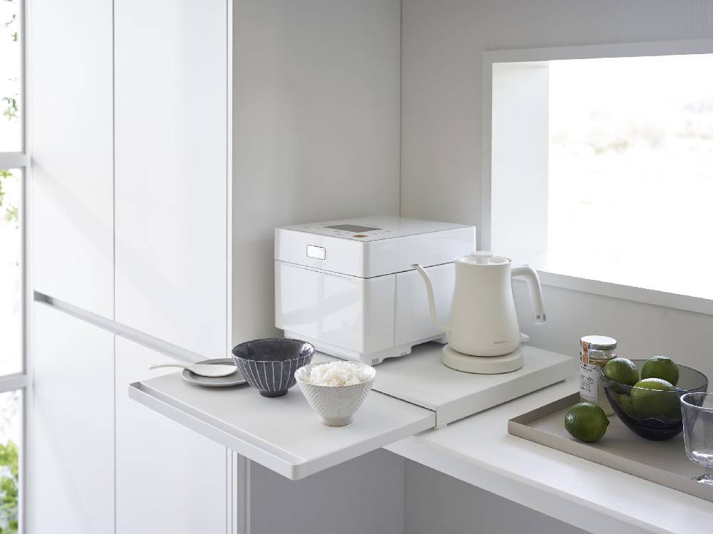 キッチンの棚下に設置されたタワー型スライドテーブル。スチール製の天板にティーポットやケトルなどの食器が並べられ、インテリアに調和する家庭用小型家電が配置されています。