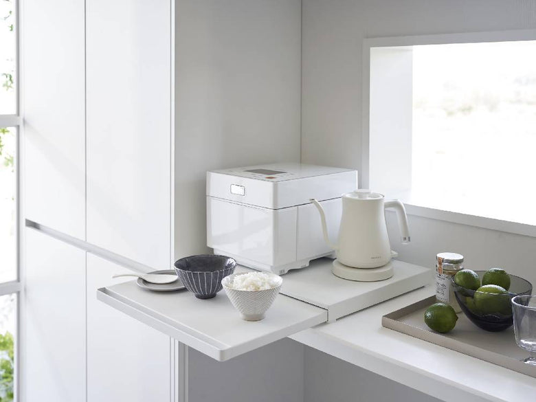 キッチンの棚下に設置されたタワー型スライドテーブル。スチール製の天板にティーポットやケトルなどの食器が並べられ、インテリアに調和する家庭用小型家電が配置されています。