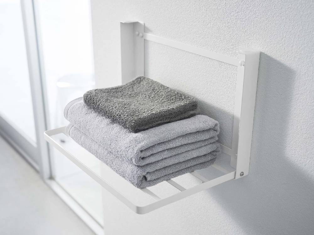 プラスチックハンドル付きスチール製タワー折り畳み棚がバスルームの石こうボード壁に設置され、タオルや日用品が収納されている様子