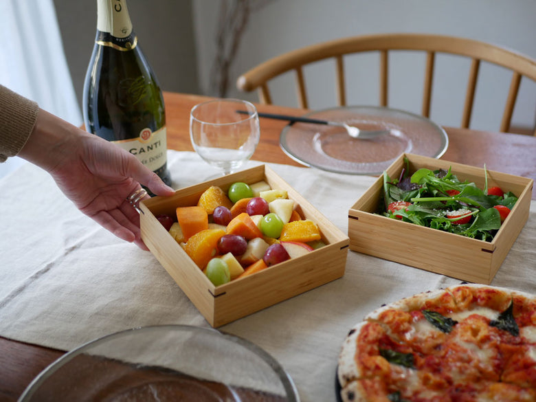 公長斎小菅の竹製二段重箱とともに、食器、果物、野菜、包丁、まな板などのキッチン用品がテーブルに美しく配置されたシーン