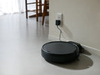 黒色のAiMYロボットクリーナーが木材の床上で自動掃除を行っている様子。電源ケーブルとブラシが見えます。