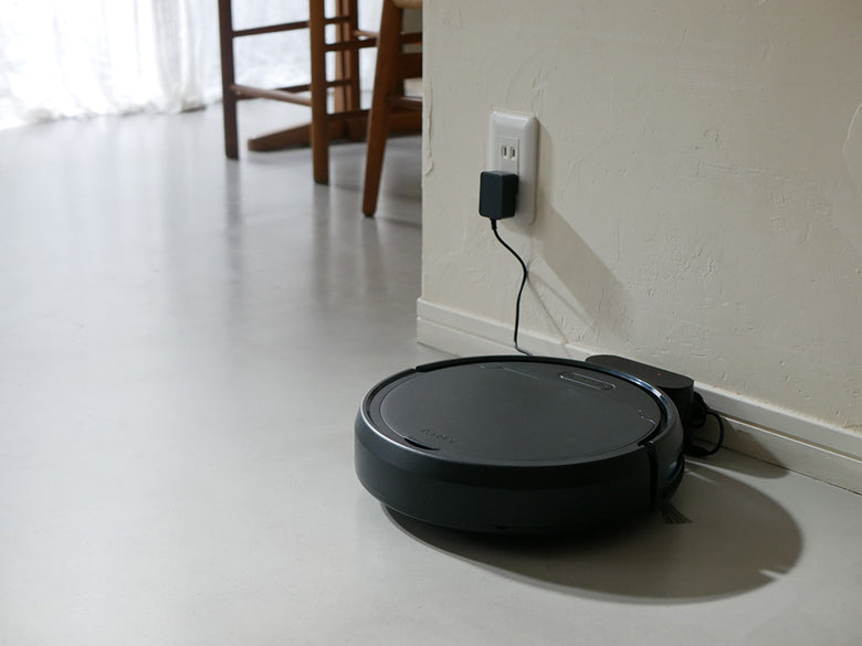 黒色のAiMYロボットクリーナーが木材の床上で自動掃除を行っている様子。電源ケーブルとブラシが見えます。