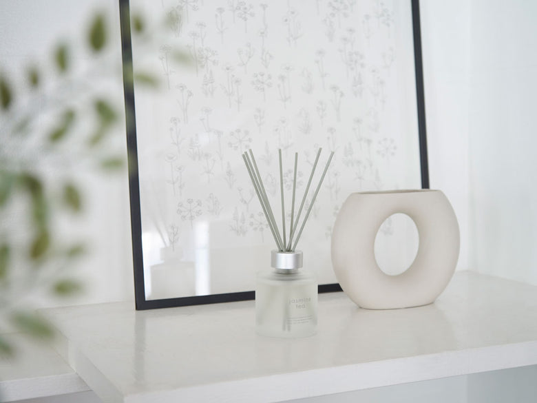 銀色の花瓶に飾られた自然素材のリードディフューザーとハンドクリームが、ランプや鉢植えと共に静物として配置された写真