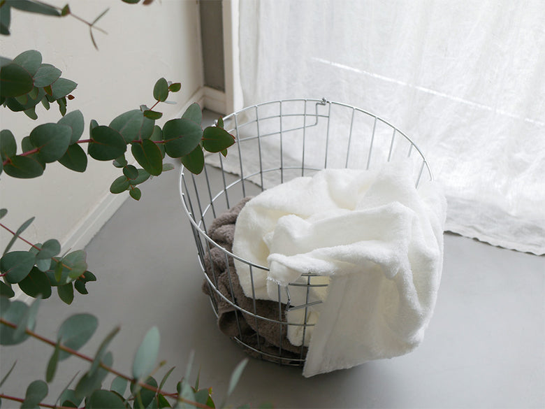 ナチュラル素材の柳編み収納バスケットにCUOLクオルのスキンケアタオルが美しく配置され、観葉植物やクッションと共に整えられた家庭用品のディスプレイ