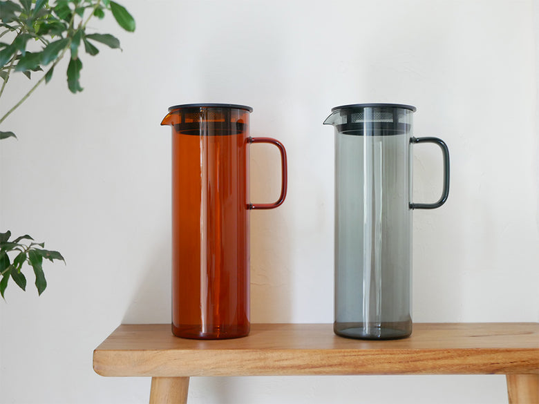 HARIOの日本製耐熱ガラス製コールドブリューピッチャーとスタックマグカップがグレーとアンバーの色合いで並べられている様子