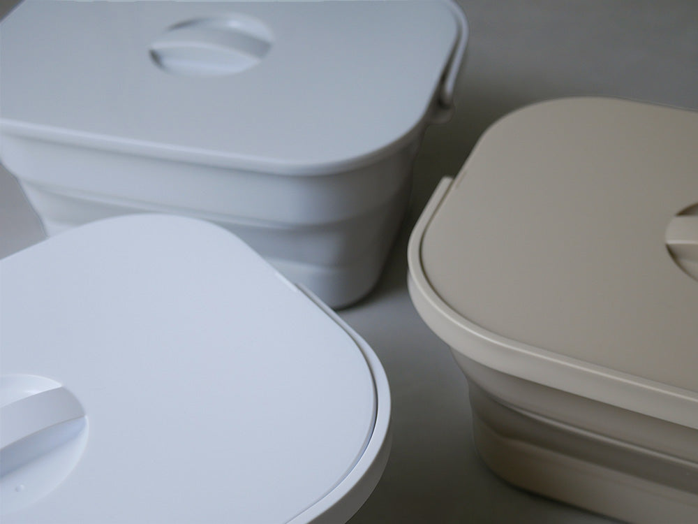 ホワイトカラーのプラスチック製折りたためるつけ置き洗いバケツ。蓋とハンドルが付いており、家庭での洗濯に便利に使用できます。