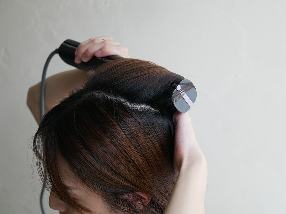 クワッズの速乾スティックドライヤーを使用して長い茶色の髪を整える女性と、ヘアケア用品が周囲に配置された写真