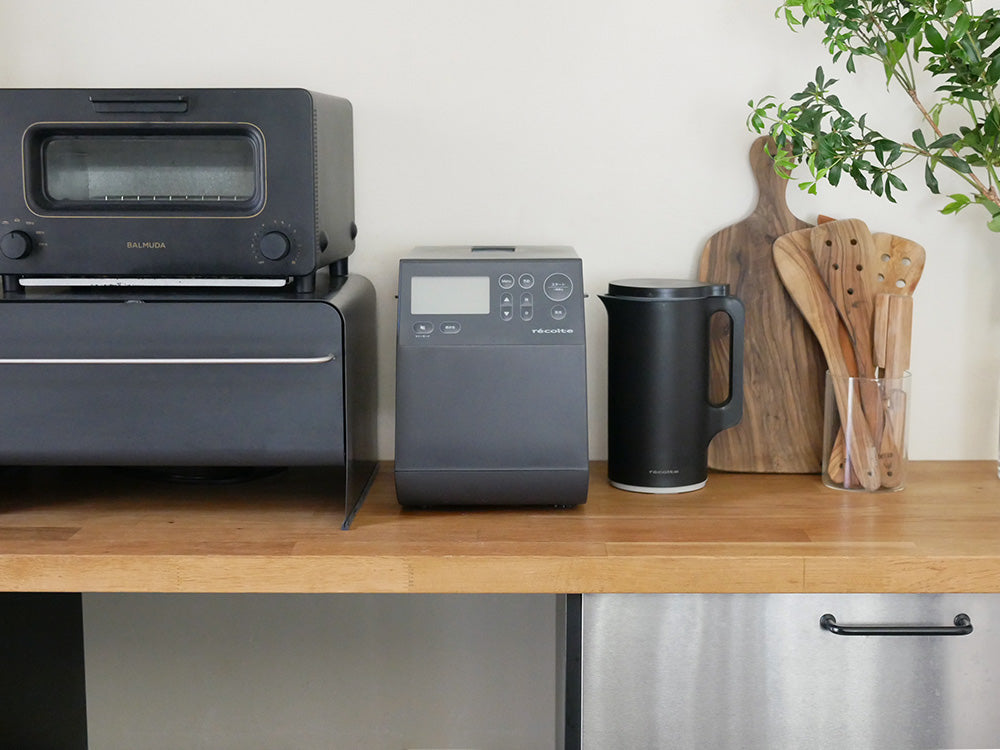 レコルトのコンパクトベーカリー本体と付属の厚釜パンケースや羽根が並んだシルバー色のキッチン家電