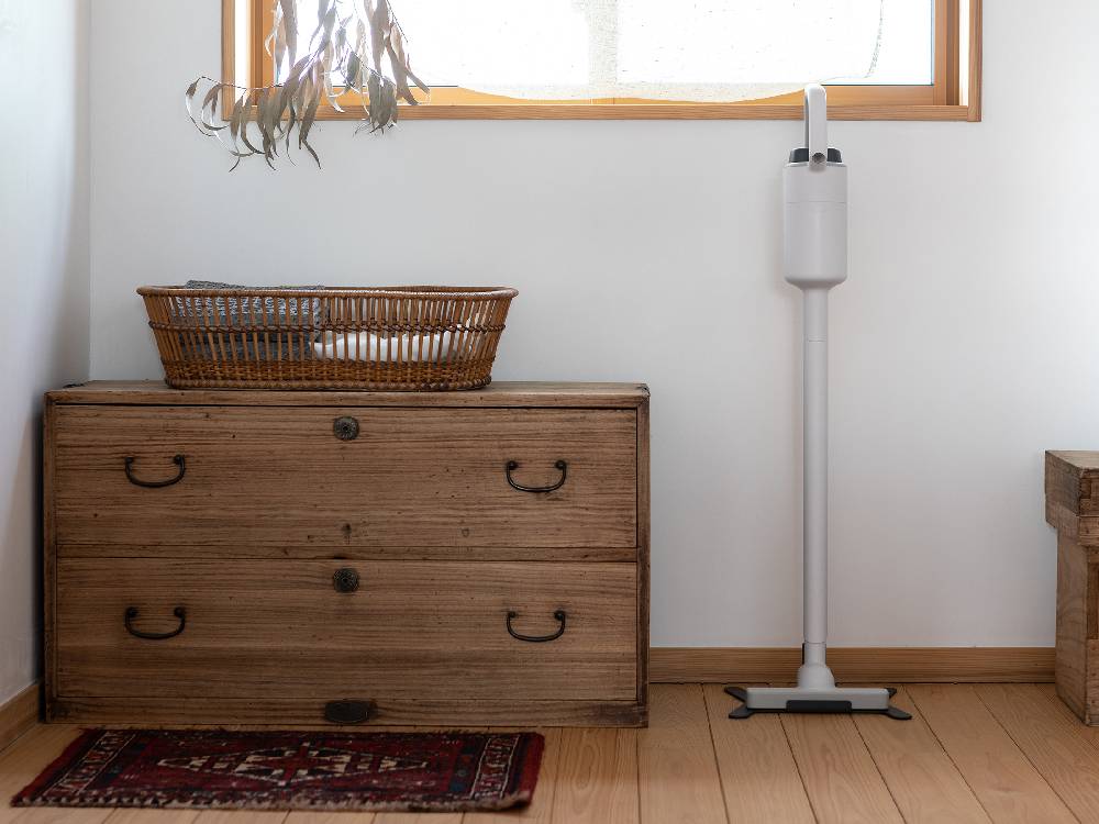 木製の床の上に置かれたプラスマイナスゼロ コードレスクリーナー G040。近くにランプと観葉植物が配置されています。