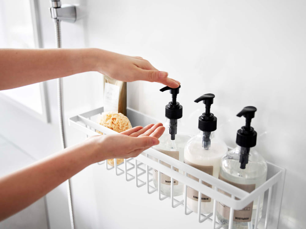 スチール製のマグネットバスルームバスケットが浴室のシンク横の棚に取り付けられ、ボトルや石鹸ディスペンサーが整然と配置されている様子