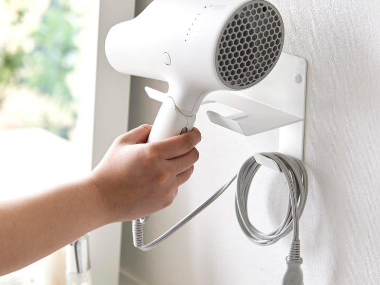 スチール製タワーウォールドライヤーホルダーが石こうボードの壁に取り付けられ、ヘアドライヤーを掛けている様子