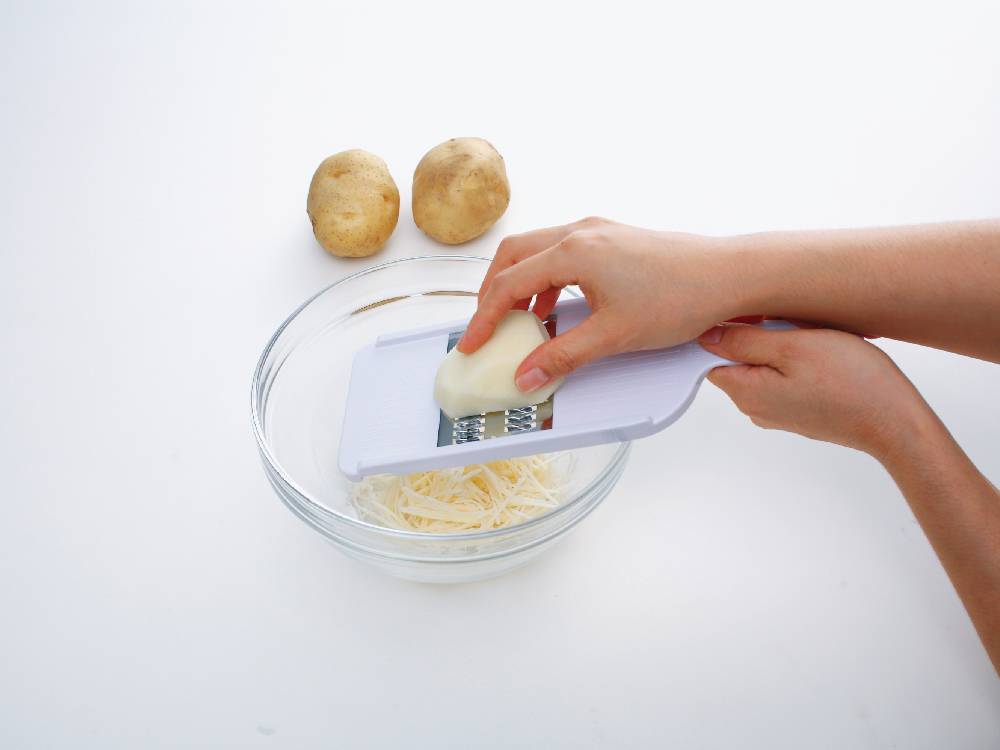 セレクト100せん切り器が切り板の上で野菜を細切りしている調理シーン。周囲にはチーズとナイフが配置されています。