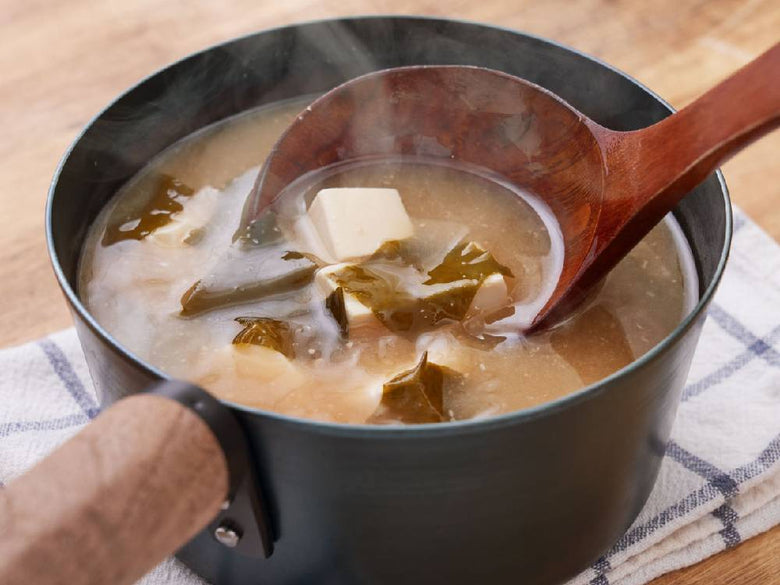 藤田金属の今日を元気にするお味噌汁パンに盛られた具だくさんの味噌汁と木製持ち手がテーブルに置かれている様子